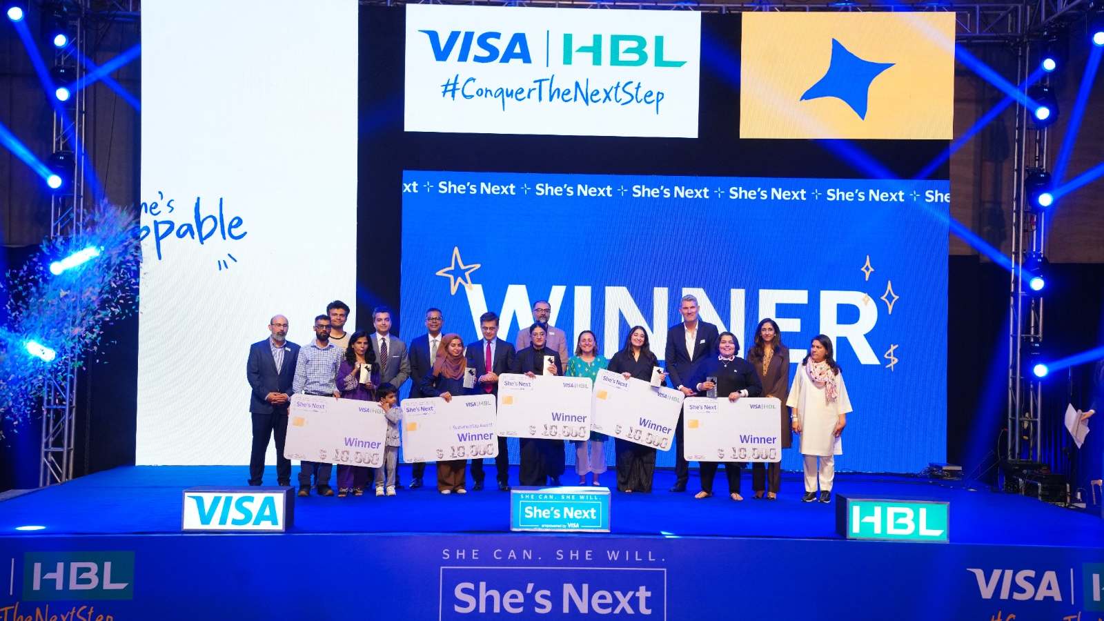 A group of people stand on a stage holding large winner checks. The backdrop features branded signage with "Winner" and "She's Next." The atmosphere is celebratory, with blue lighting and a dynamic design conveying achievement and success.